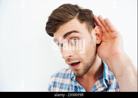 Ich will alles wissen! Neugieriger junger Mann, der Hand hinter Ohr hält und in die Kamera blickt, während er vor weißem Hintergrund steht Stockfoto