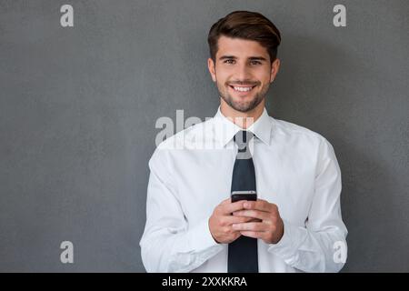 Mit meinen Kollegen in Kontakt bleiben. Hübscher junger Mann in Hemd und Krawatte, der Handy auslöscht und lächelt, während er vor grauem Hintergrund steht Stockfoto