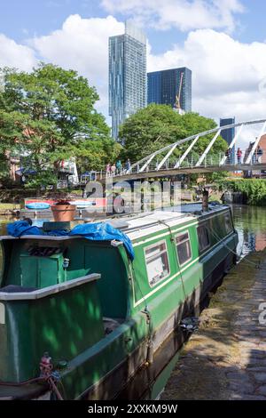 Manchester, Greater Manchester, Großbritannien. 24. August 2024: Ein grünes schmales Boot liegt an einem friedlichen Kanal, umgeben von einem üppigen grünen Baum und modernem Hi Stockfoto