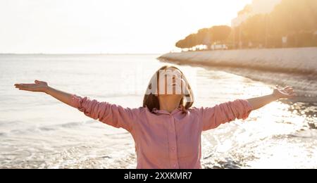 Frau genießt freudig den Sonnenuntergang am Strand mit ausgestreckten Armen Stockfoto