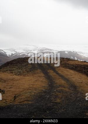 Eine abgeschiedene Schotterstraße schlängelt sich entlang eines Berges, flankiert von verschneiten Berggipfeln, die einen dramatischen und isolierten Pfad durch das zerklüftete Gelände schaffen. Stockfoto