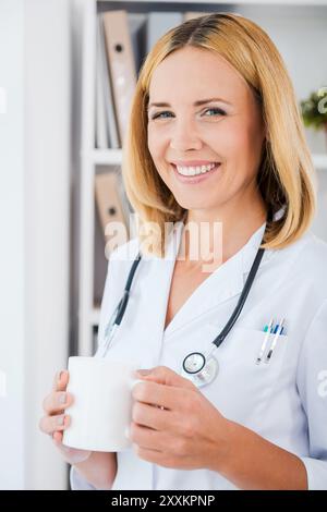 Pause von einer schweren Arbeit. Fröhliche Ärztin in weißer Uniform mit Tasse und Blick in die Kamera Stockfoto