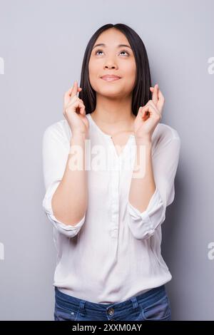 Warten auf besondere Momente. Fröhliche junge Frau, die aufblickt und die Daumen gedrückt hält, während sie vor grauem Hintergrund steht Stockfoto