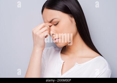 Depressive Frau. Depressive junge asiatische Frau, die ihren Kopf mit den Händen berührt und die Augen geschlossen hält, während sie vor grauem Hintergrund steht Stockfoto