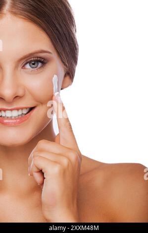Hautpflege. Zugeschnittenes Bild einer jungen Frau, die Creme auf ihrem Gesicht spreizt und lächelt, während sie vor weißem Hintergrund steht Stockfoto