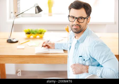 Auf Erfolg ausgerichtet. Hübscher junger Mann in Hemd und Brille, der in den Notizblock schreibt und die Kamera über den Schultern ansieht, während er an seiner Arbeitsplatte sitzt Stockfoto