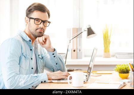 Neue Vision schaffen. Hübscher junger Mann in Hemd und Brille, der am Laptop arbeitet und die Kamera ansieht, während er an seinem Arbeitsplatz sitzt Stockfoto