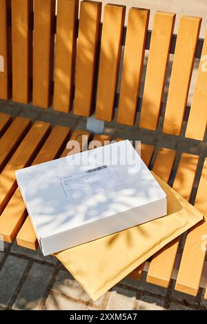 Weißes Paket und gelber gepolsterter Umschlag, der unter direktem Sonnenlicht auf einer Holzbank platziert wird und Schatten der Bank zeigt Stockfoto