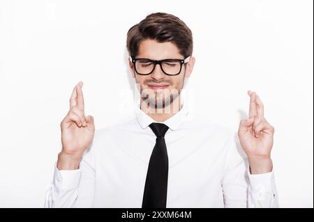 Warten auf besondere Momente. Junger Mann, der vor weißem Hintergrund die Daumen drückt und die Augen geschlossen hat Stockfoto