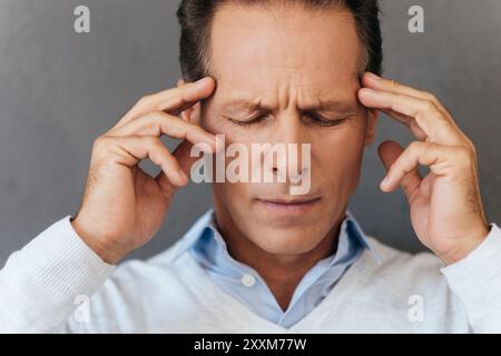 Schreckliche Kopfschmerzen. Frustrierter reifer Mann, der seinen Kopf mit Fingern berührt und die Augen geschlossen hält, während er vor grauem Hintergrund steht Stockfoto