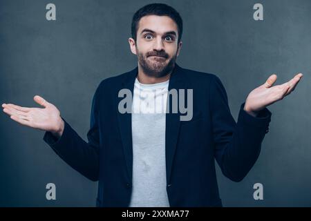 Wer weiß? Verwirrter junger Mann, der seine Arme ausstreckt und grimmig vor grauem Hintergrund steht Stockfoto