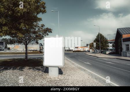 Eine leere Plakatwand steht in einer ruhigen Stadtstraße, umgeben von Gebäuden, Bäumen und einem Kreisverkehr. Der leere Anzeigenbereich ist bereit für benutzerdefinierte Werbung Stockfoto