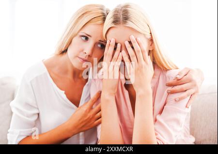 Ich werde immer bei dir sein, in jeder Situation! Schöne reife Frau tröstet ihre weinende Tochter, während sie auf dem Sofa sitzt Stockfoto