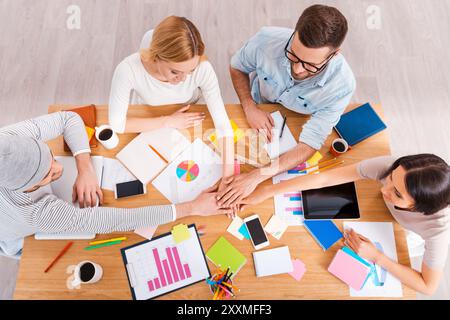 Das beste kreative Team. Blick von oben auf eine Gruppe von Geschäftsleuten in eleganter Freizeitkleidung, die die Hände zusammenhalten, während sie am Holztisch sitzen Stockfoto