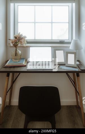 Ein minimalistisches Home Office-Setup mit einem Laptop mit leerem Bildschirm, einer Vase mit Blumen und zwei leeren Bilderrahmen auf einem Block Stockfoto