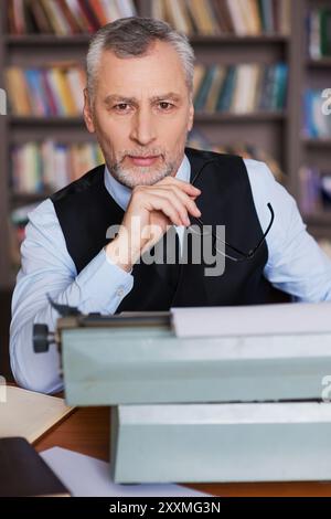 Gerade inspiriert. Selbstbewusster, grauer Seniorenmann in formaler Kleidung, der an der Schreibmaschine sitzt und in die Kamera mit Bücherregal im Hintergrund blickt Stockfoto