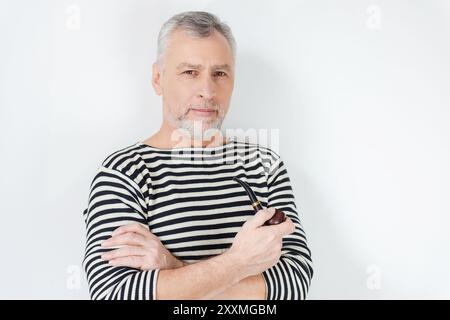 Souveräner Seemann. Selbstbewusster, älterer bärtiger Mann in gestreifter Kleidung, der eine Pfeife raucht und in die Kamera blickt, während er vor weißem Hintergrund steht Stockfoto