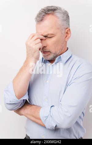 Müde und gestresst. Frustrierter Senior Mann im Hemd, der den Kopf mit den Fingern berührt und die Augen geschlossen hält, während er vor weißem Hintergrund steht Stockfoto