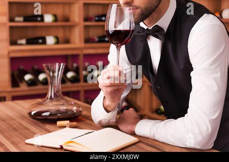 Sommelier untersucht Wein. Zugeschnittenes Bild eines selbstbewussten männlichen Sommeliers, der Wein untersucht, während er ihn riecht und sich an den Holztisch mit Weinregal i lehnt Stockfoto