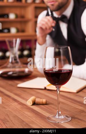 Schreiben von Notizen über Wein. Zugeschnittenes Bild eines nachdenklichen männlichen Sommeliers, der sein Kinn mit Stift berührt, während er sich an den Holztisch lehnt, mit Weinglas an der Th Stockfoto