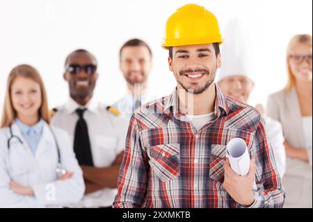 Wenn ich groß bin, werde ich Ingenieur. Selbstbewusster junger Mann mit Hardhat, der einen Plan hält und lächelt, während eine Gruppe von Menschen in verschiedenen Berufen stan Stockfoto