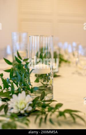 Glaszylindervase in der Mitte mit Wasser gefüllt und mit einer schwebenden weißen Kerze auf dem Tisch mit grünen Blättern Stockfoto