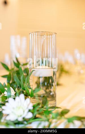 Glaszylindervase Mittelstück gefüllt mit Wasser und mit einer schwimmenden weißen Kerze auf Tisch mit grünen Blättern bei Hochzeitsempfang Stockfoto