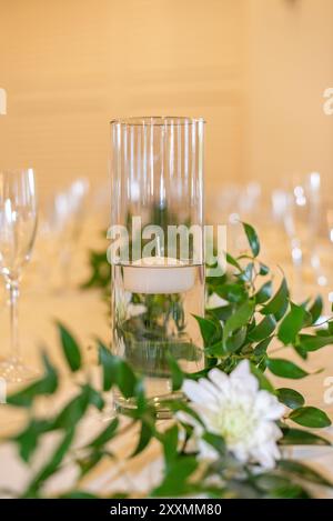 Glaszylindervase in der Mitte, gefüllt mit Wasser und mit einer schwimmenden weißen Kerze auf dem Tisch mit grünen Blättern im Vordergrund Stockfoto