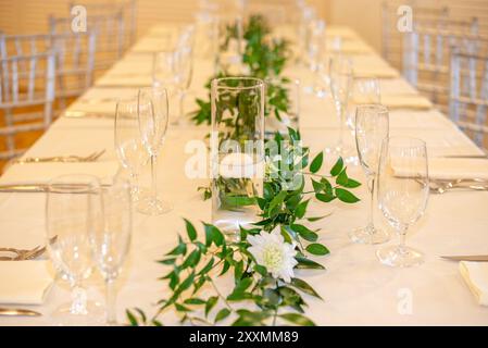 Glaszylindervase in der Mitte, gefüllt mit Wasser und mit einer schwimmenden weißen Kerze auf dem Tisch, umgeben von Glaswaren und grünen Blättern Stockfoto