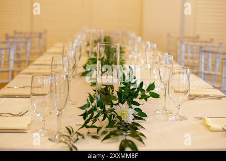 Glaszylindervase in der Mitte, gefüllt mit Wasser und mit einer schwimmenden weißen Kerze auf dem Tisch, umgeben von Glaswaren und grünen Blättern Stockfoto