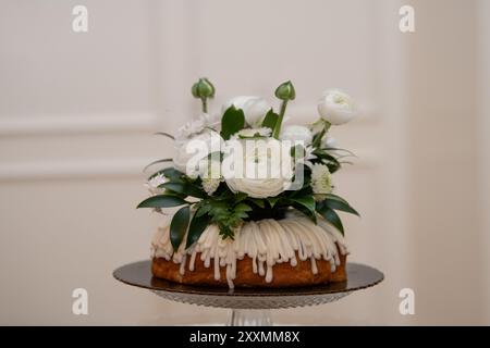 Vanilatekuchen mit Vanilleglasur, dekoriert mit weißen Rosen, Gänseblümchen und Grün auf Glaskuchensockel mit weißem Wandhintergrund Stockfoto