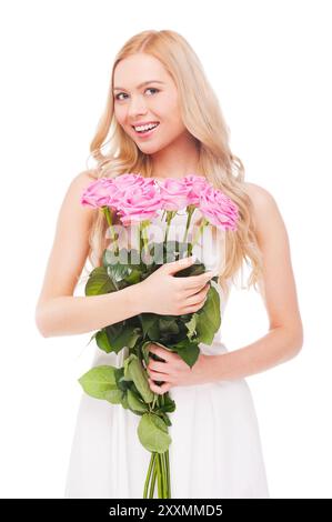 Es ist ein großer Tag für mich! Schöne junge Frau mit blondem Haar, die einen Blumenstrauß mit rosa Rosen hält und lächelt, während sie isoliert auf weiß steht Stockfoto