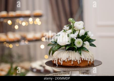 Vanillegebäck mit Vanilleglasur, dekoriert mit weißen Rosen, Gänseblümchen und Grün auf Glaskuchensockel mit verschwommenem Cupcake-Hintergrund Stockfoto