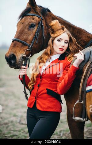 Rothaarige Jockey-Mädchen in einer roten Strickjacke und schwarzen hohen Stiefeln mit einem Pferd für einen Spaziergang Stockfoto