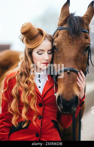 Rothaarige Jockey-Mädchen in einer roten Strickjacke und schwarzen hohen Stiefeln mit einem Pferd für einen Spaziergang Stockfoto