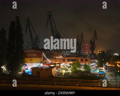 Danzig, Polen - 13. Juli 2024: Nächtliche Stadtlandschaft mit beleuchteten Gebäuden und hoch aufragenden Kränen im Vordergrund, die das geschäftige Treiben der Stadt zeigen Stockfoto