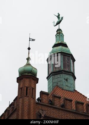 Danzig, Polen - 13. Juli 2024: Eine wunderschöne architektonische Aufnahme, die einen historischen grünen Kupferturm und ein detailliertes Backsteingebäude mit komplizierten St. Stockfoto