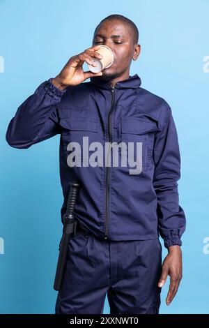 afroamerikanischer Agent trinkt eine Kaffeetasse vor blauem Hintergrund und serviert Koffeinerfrischungen, um nach einer schlaflosen Nacht mehr Energie zu bekommen. Männlicher Wachmann posiert vor der Kamera. Stockfoto