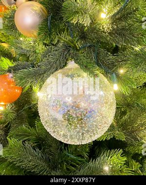 Wunderschöne, strukturierte Glaskugel-Ornamente in Nahaufnahme, die an grünen Nadelblättern mit kleinen Lichtern, goldenen und orangen Ornamenten hängen Stockfoto