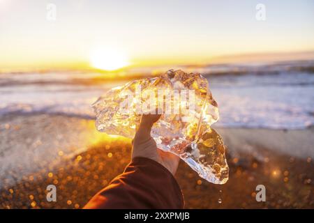 Ein Mann hält ein Stück Eis in der Hand bei Sonnenaufgang am Diamond Beach neben Jokulsarlon im Winter in Island Stockfoto