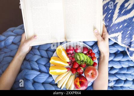 Mit frischem Obstsalat Platte auf einem Blue Plaid und Buch. Frau liest das Buch und das Essen von frischem Obst. Close Up. Top Ausblick. Gemütlich und gesund Konzept Stockfoto