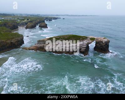 Ein markanter Felsen in Form einer natürlichen Brücke, umgeben von schäumenden Wellen und einer verlockenden Küste, aus der Vogelperspektive, Praia das Catedrais, Playa de Stockfoto