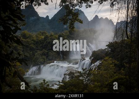 Neblige Wasserfälle stürzen bewaldete Klippen hinunter vor der Kulisse von hoch aufragenden Bergen, die von dichtem Laub unter einem bewölkten Himmel eingerahmt werden Stockfoto