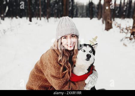 Junge schöne glücklich lächelnde Frau in Pelz umarmenden Husky Hund in roten Weihnachtsschal und festliche Hirschhörner gekleidet. Portrait Foto von Mädchen und tun Stockfoto