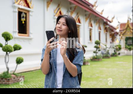 Eine schöne, positive junge asiatische Touristin in einer Jeansjacke schlendert mit einem Smartphone in der Hand durch einen Tempel und genießt ihren Besuch in Thaila Stockfoto