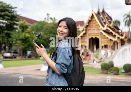 Eine schöne, positive junge asiatische Touristin in einer Jeansjacke schlendert mit einem Smartphone in der Hand durch einen Tempel und genießt ihren Besuch in Thaila Stockfoto