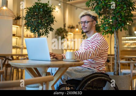 Ein fokussierter Mann mit Behinderungen im Rollstuhl, der online mit einem Laptop arbeitet, sitzt in einem gemütlichen Café Stockfoto