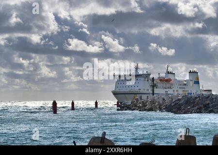 Eine große Fähre fährt durch einen Hafen, der durch dramatischen, bewölkten Himmel und einen Wellenbrecher hervorgehoben wird, der den Transport und die Infrastruktur des Meeres veranschaulicht Stockfoto