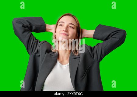 Frau in Black Blazer lehnt sich mit den Händen hinter Kopf gegen Green Screen zurück Stockfoto