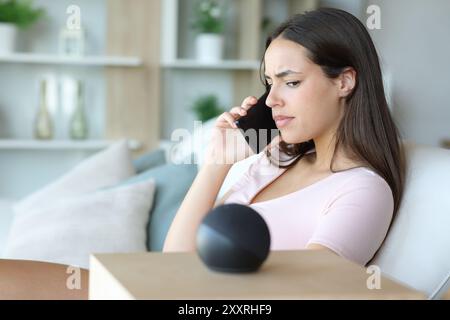 Verdächtige Frau, die am Telefon spricht und zu Hause auf intelligente Lautsprecher schaut Stockfoto